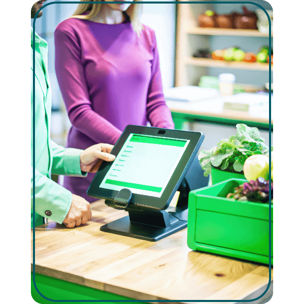 Why Choose Paywint for Your Point of Sale System? A Modern Point of Sale System with a Touch-Screen Interface Displayed on a Countertop, Surrounded by Fresh Produce in a Grocery Setting. Two Individuals Are Interacting near the System, Showcasing a Seamless Checkout Process