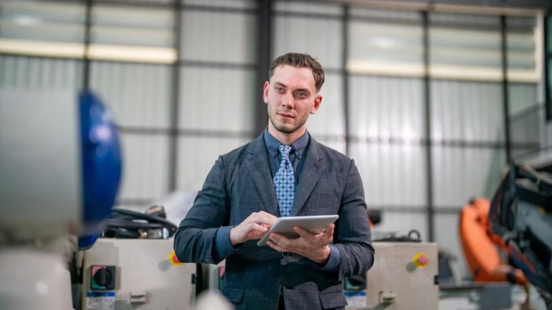 A Person in a Suit Holds a Tablet, Standing in a Well-Lit Industrial Workspace with Machinery in the Background
