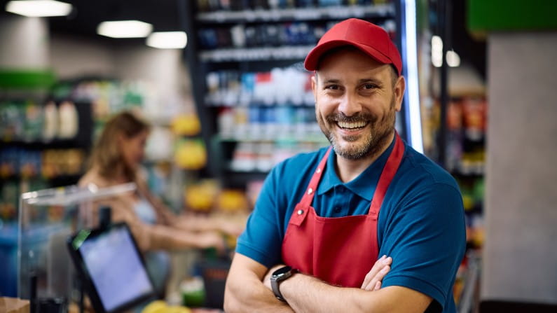 A Cashier Wearing a Red Apron and Cap Stands Confidently with Arms Crossed in a Well-Stocked Convenience Store